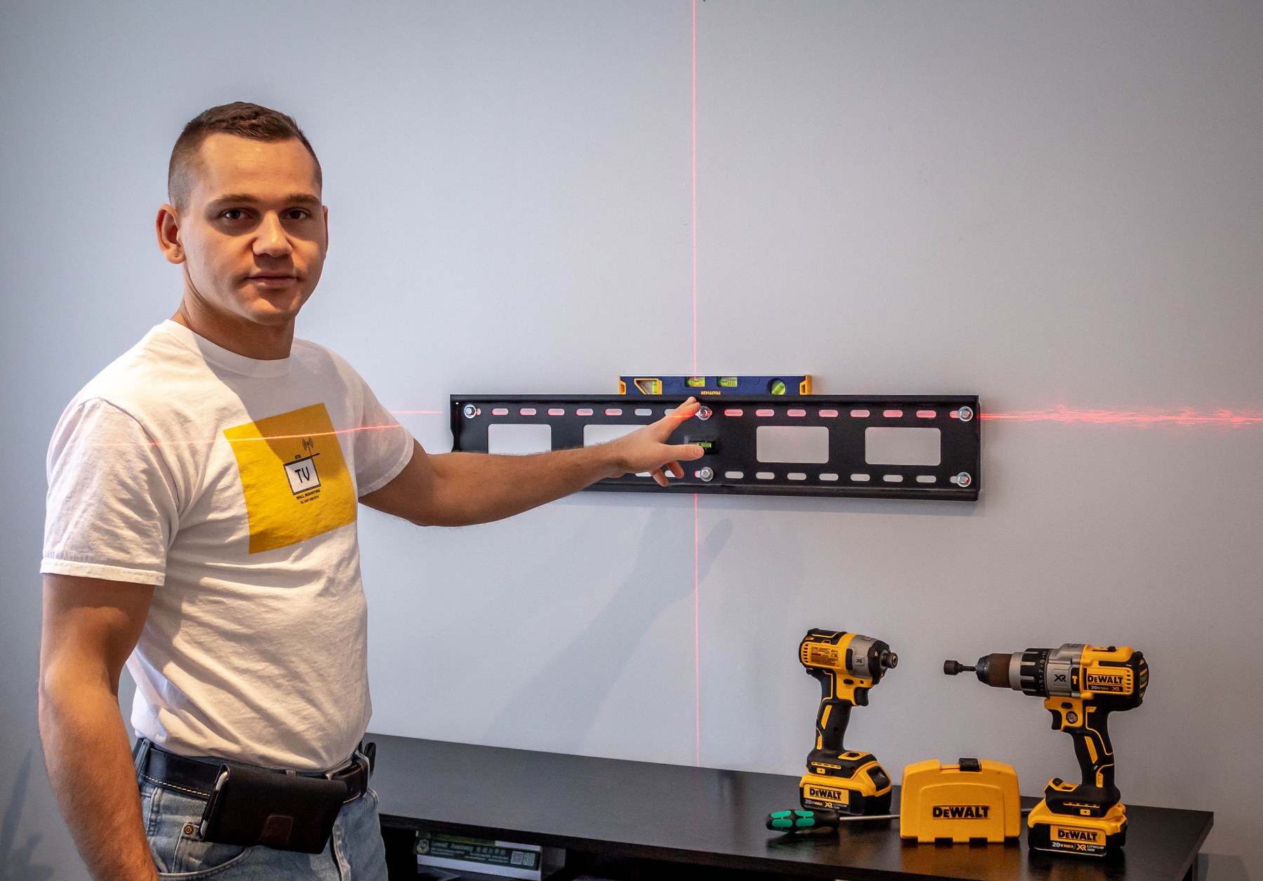 GTA TV Wall Mounting technician setting a tilting TV bracket to a cross-line laser, with an Irwin box level across the rail and DeWalt drivers on the console below