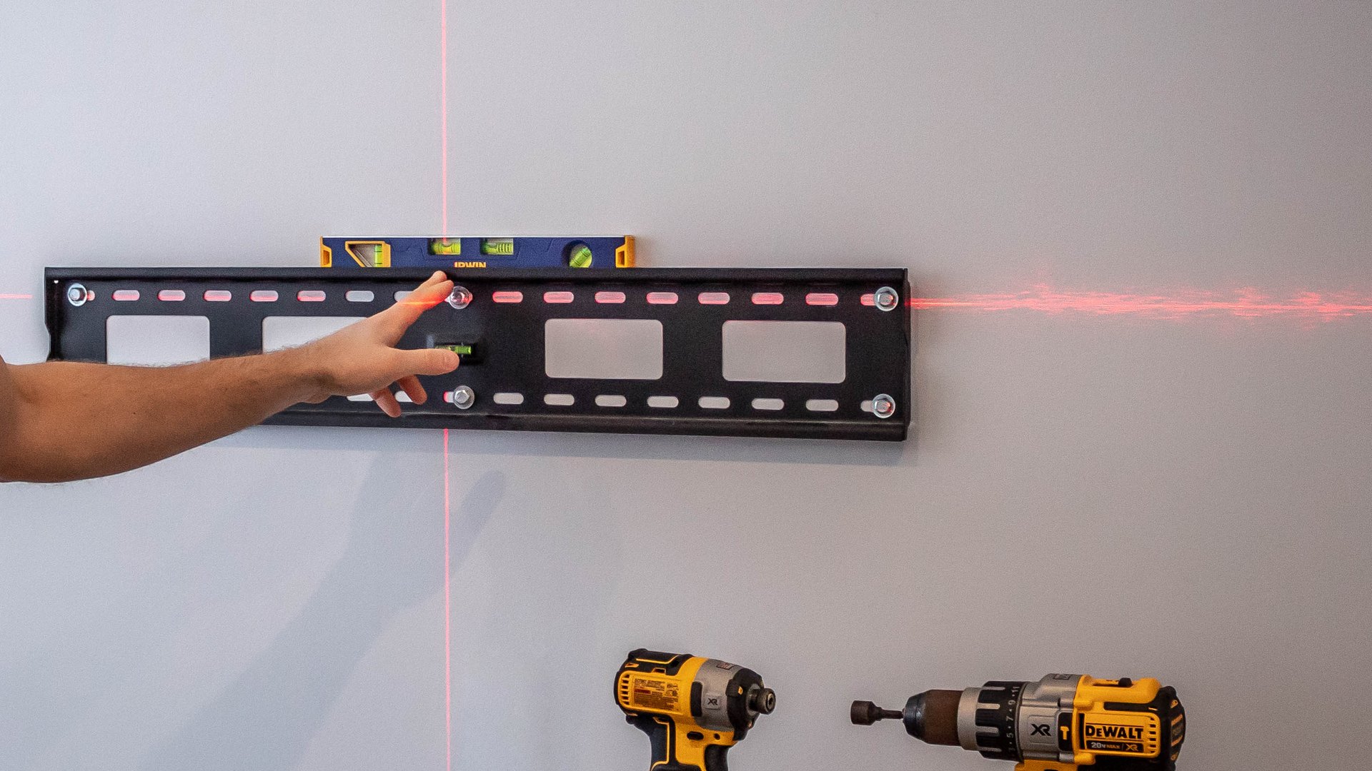 A GTA TV Wall Mounting technician levelling a wall-mount bracket with a cross-line laser, with two drills staged and ready before any hole is drilled