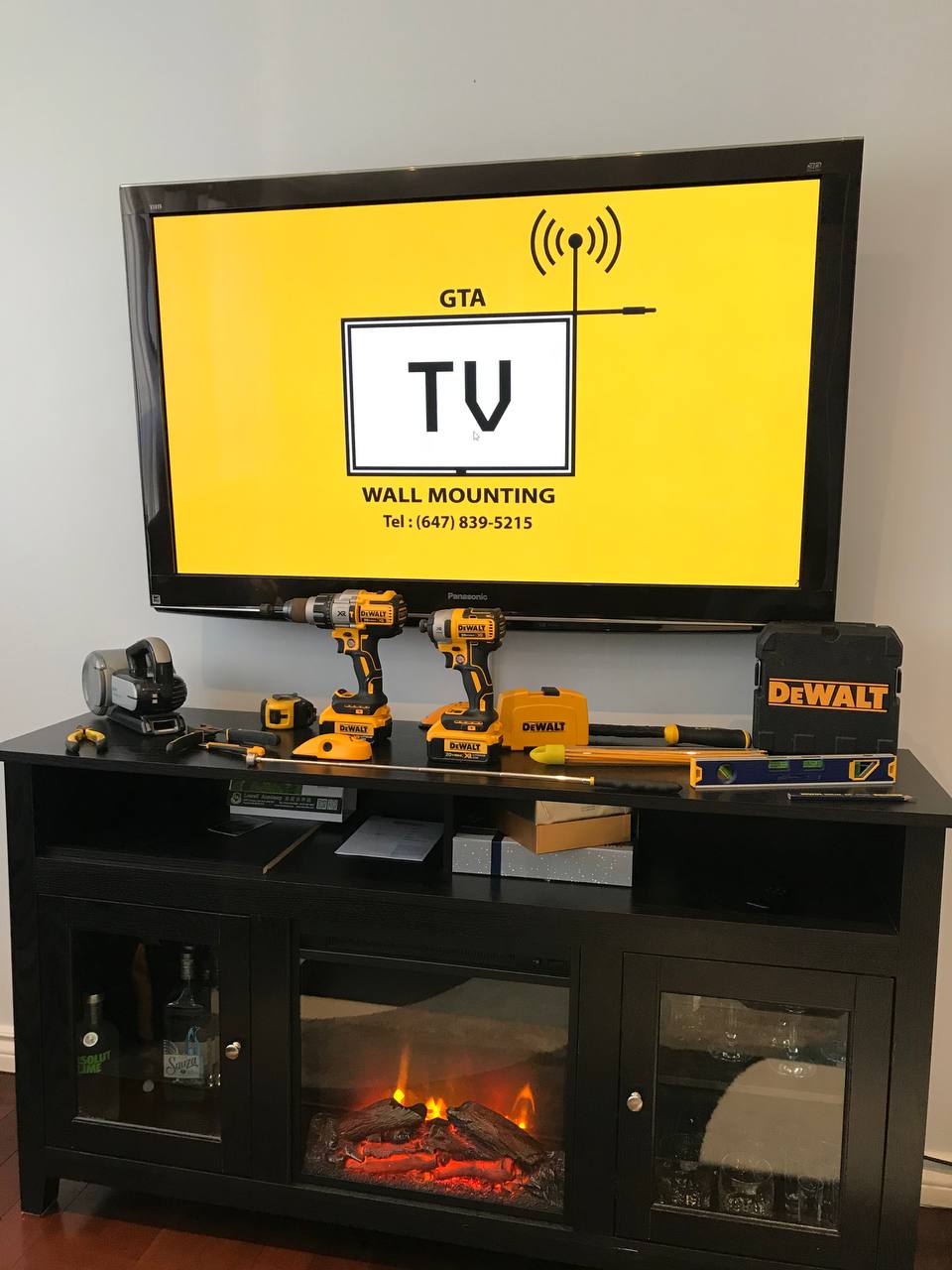 The GTA TV Wall Mounting tool kit laid out below a mounted Panasonic screen: hammer drill, impact driver, laser, levels and masonry bits