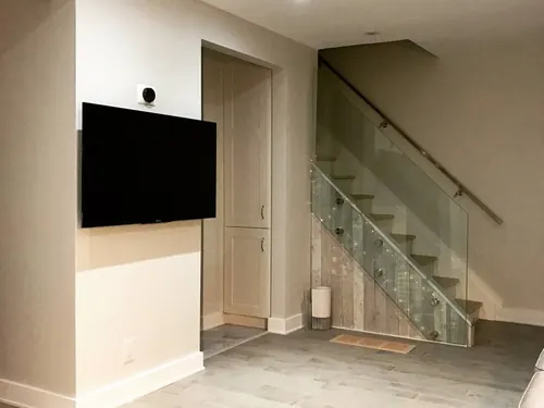 TV mounted on a basement half-wall beside a glass-railed staircase, with a sensor fitted above the screen