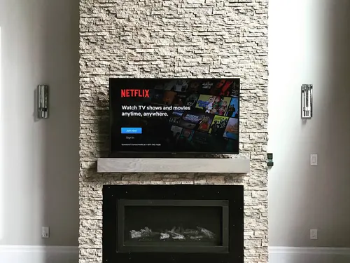 TV mounted on a stacked stone chimney above a timber mantel and linear fireplace, between two sconces