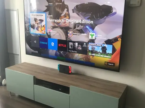 Large TV mounted on a white wall above a sage green and oak console with a games console below