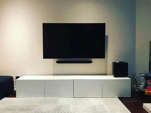 TV and soundbar mounted on a cream wall above a white console, with a subwoofer alongside