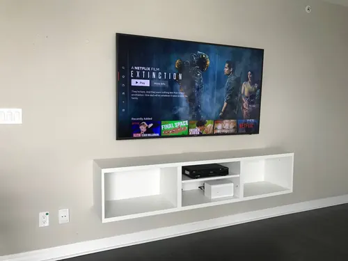 TV mounted on a beige wall above a white floating media cubby, with the outlet and cable plates alongside