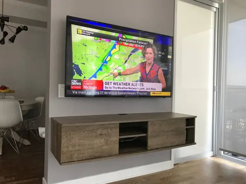 TV mounted on an angled condo wall above a grey floating console, with the dining area beyond