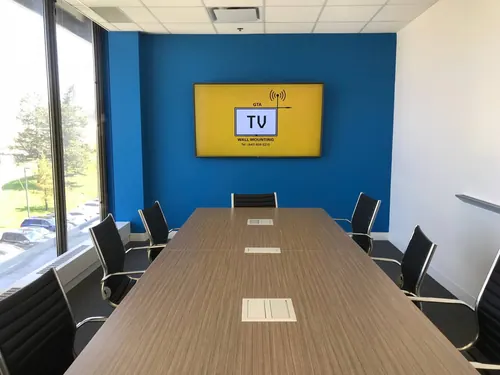 TV mounted on a blue boardroom wall, seen down the length of the table toward the window