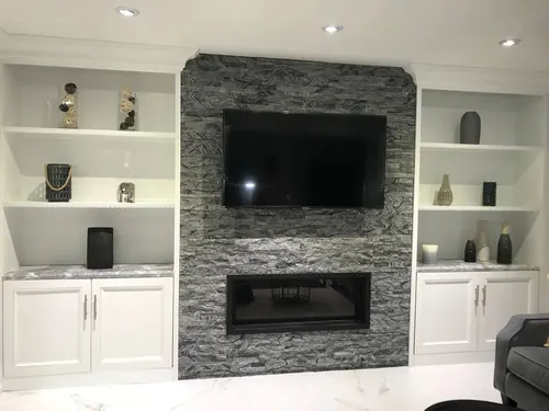 TV mounted on a grey stacked stone chimney above a linear fireplace, between marble-topped built-ins
