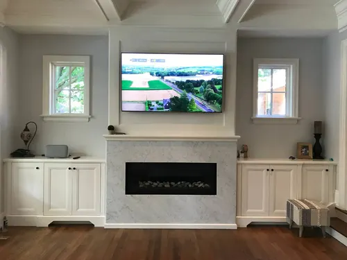 TV mounted above a marble fireplace surround and painted mantel, between white cabinets and two windows