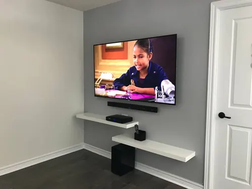 TV and soundbar mounted on a grey wall above two staggered white floating shelves and a subwoofer