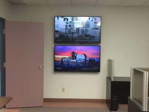 Two TVs stacked one above the other on an office wall beside workstation partitions