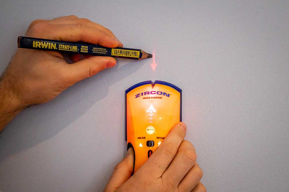Installer sweeping a wall with an electronic stud detector to confirm stud positions before marking a TV bracket