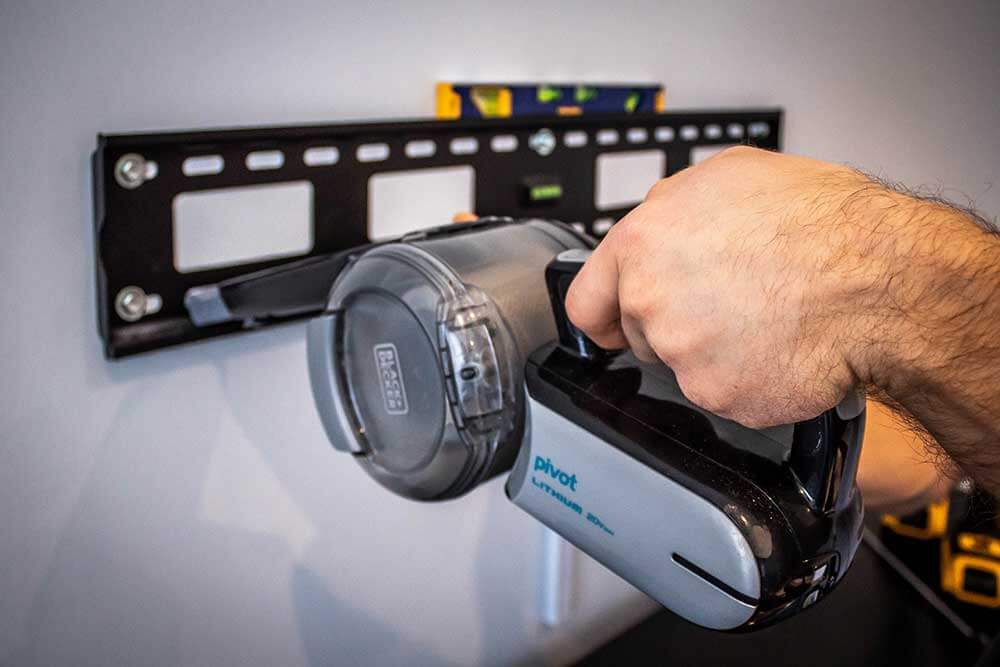 Installer drilling a TV bracket fixing while holding a vacuum nozzle at the bit to catch the dust
