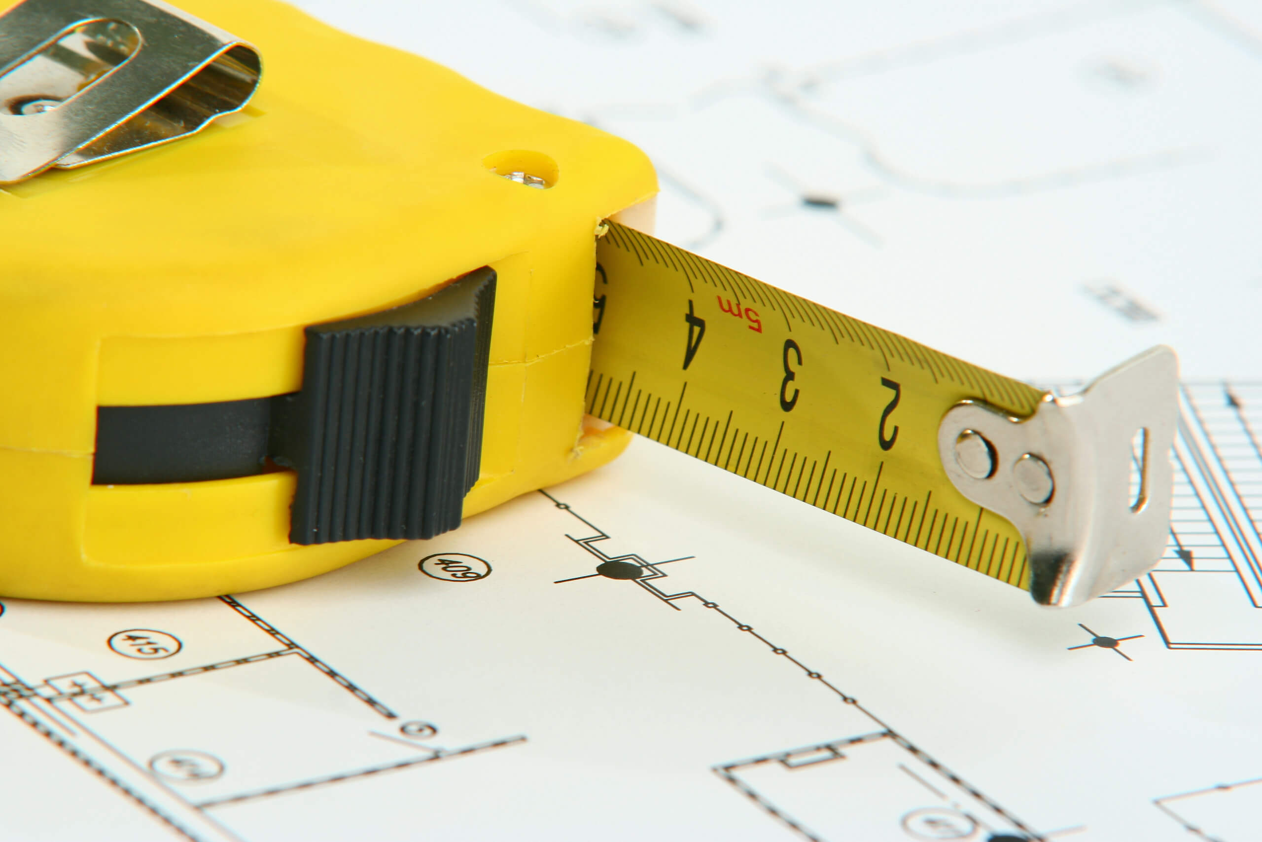 A close-up of a yellow tape measure extended over a set of architectural blueprints, perhaps for planning TV wall mounting. The tape measure shows measurements in both inches and centimeters. The end of the tape is slightly worn, and the blueprints display various lines and symbols for a building layout.