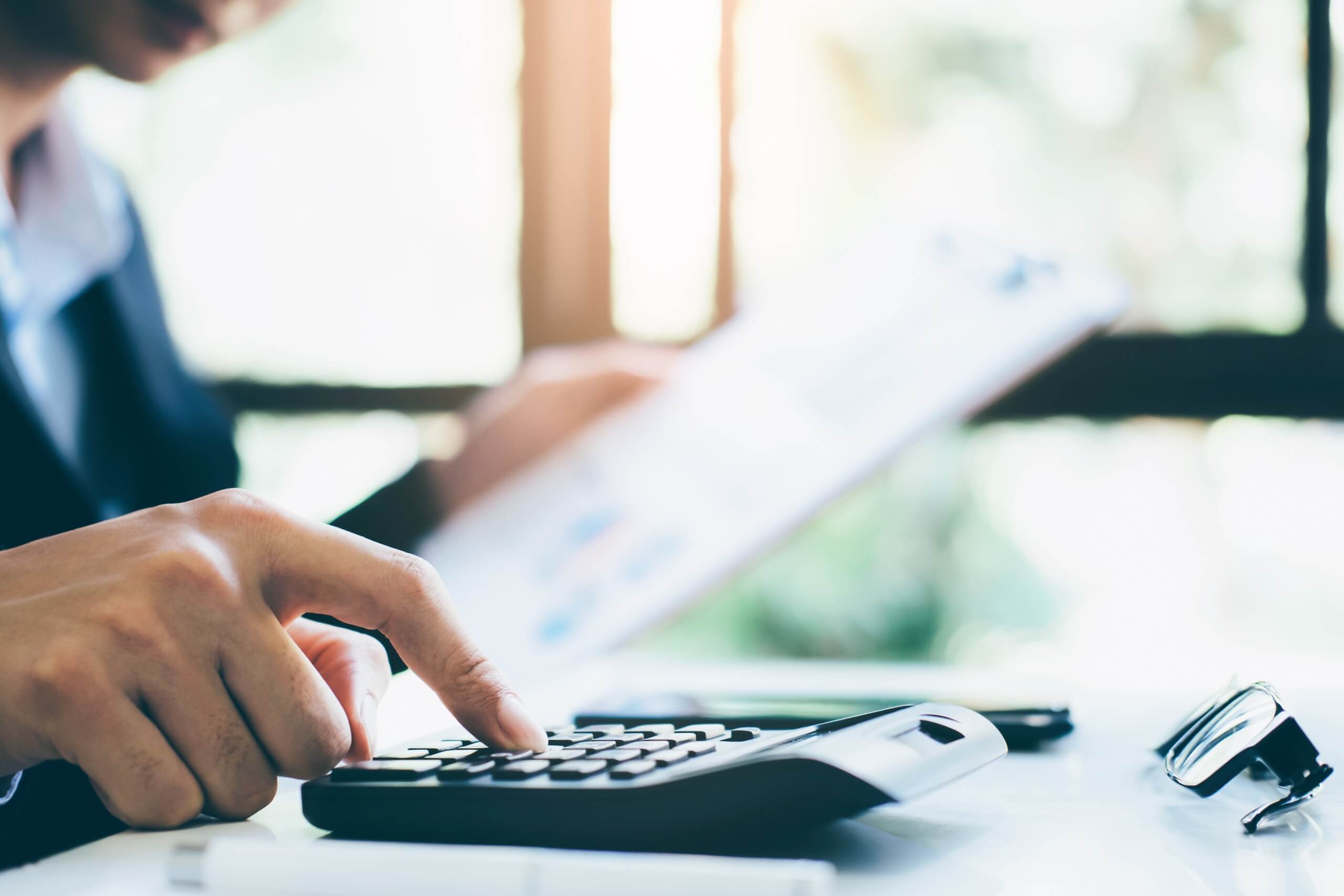 A person pressing buttons on a calculator at a desk while holding a document in the other hand. In the background, there is a window letting in sunlight, and a pair of eyeglasses is placed on the desk beside the calculator. Beside the desk, you can see tools used for TV wall mounting. The focus is on the hand and calculator.