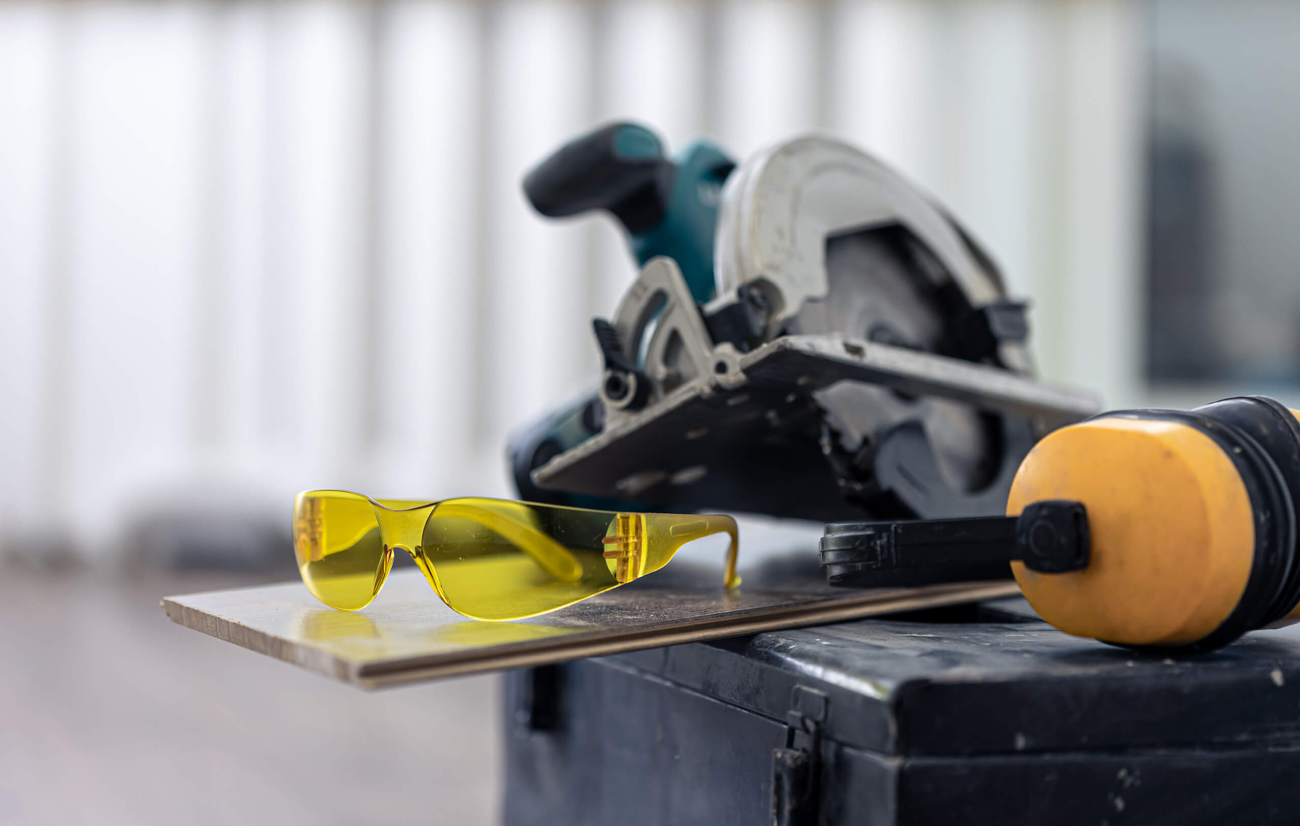 Yellow safety glasses resting on a board, with a circular saw and ear protection behind them.