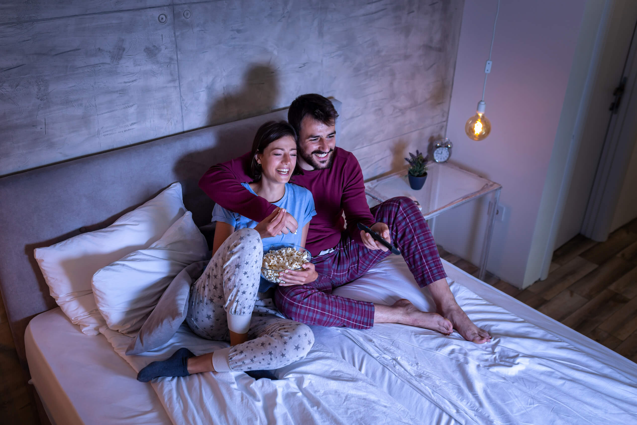 A couple is sitting on a bed in cozy pajamas, smiling and holding a bowl of popcorn. The man is pointing a remote control at their TV wall mounting off-screen. The room is softly lit, creating a warm and inviting atmosphere.