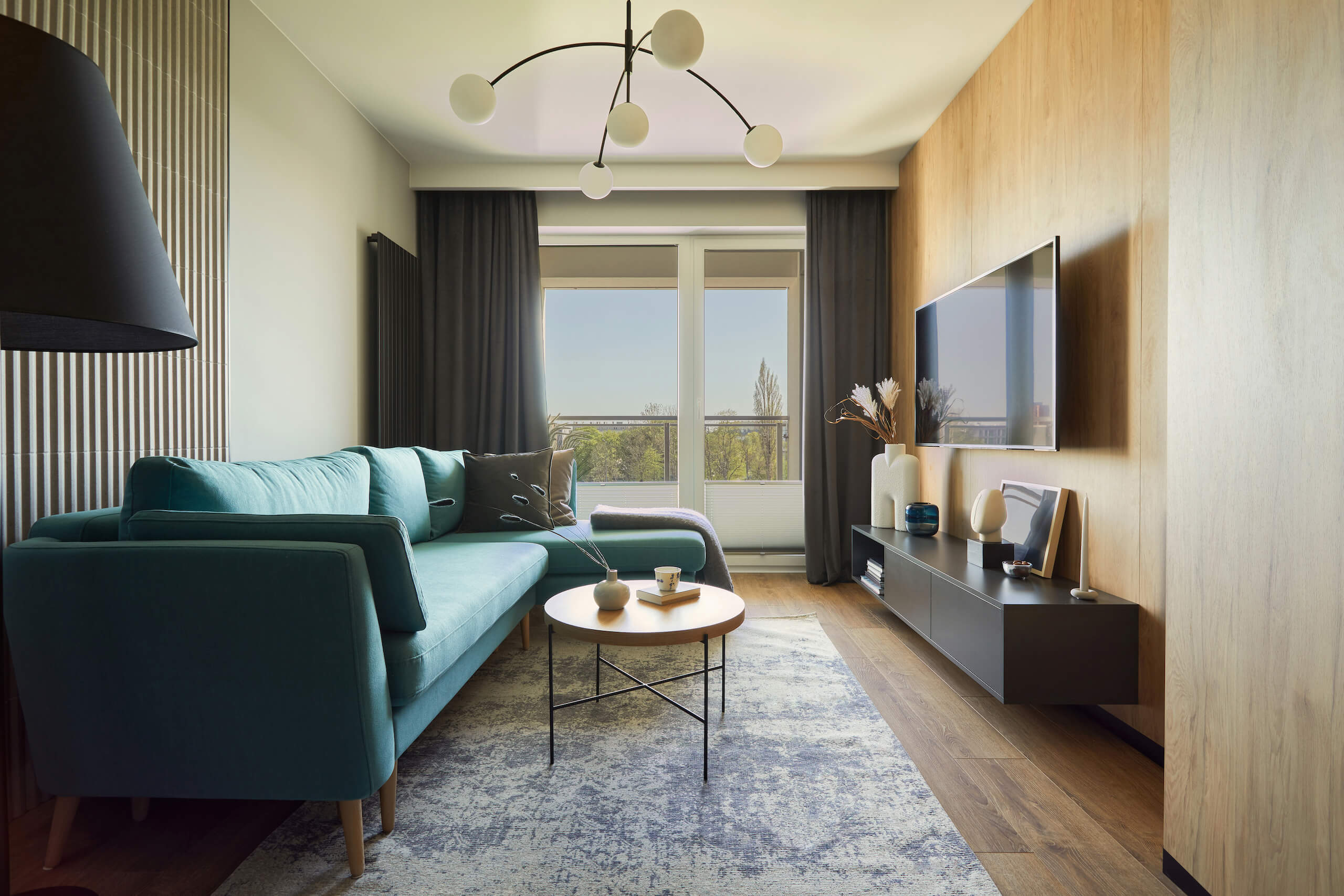 A modern living room with a teal sofa, round wooden coffee table, and TV wall mounting on a wooden accent wall. Neutral-toned rug on a wooden floor, large window with gray curtains, decorative shelves, a black floor lamp, potted plants, and abstract chandelier.