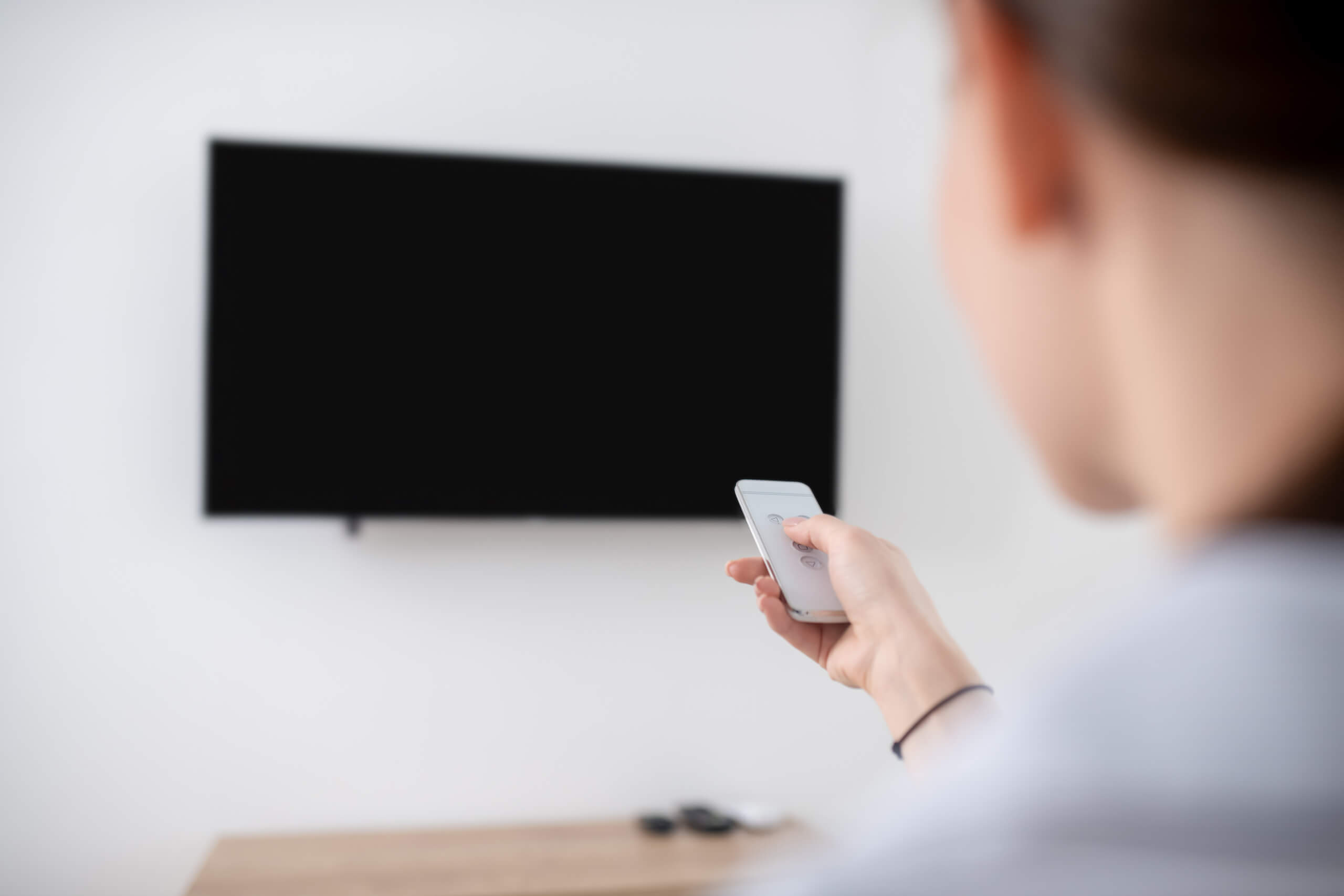 A person holding a remote control, pointing it at a blank, wall-mounted television screen in a minimalistic room. The image is taken from behind and focuses on the hand and remote, highlighting the sleek design of the TV wall mounting in the distance.