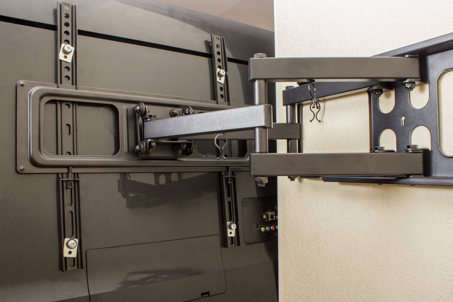 A close-up image shows a modern wall-mounted TV bracket fixed to a flat-screen television. The mount has a multi-joint arm, allowing it to extend and swivel, enabling flexible TV positioning. The background features a light-colored wall with a subtle textured pattern, showcasing expert TV wall mounting.
