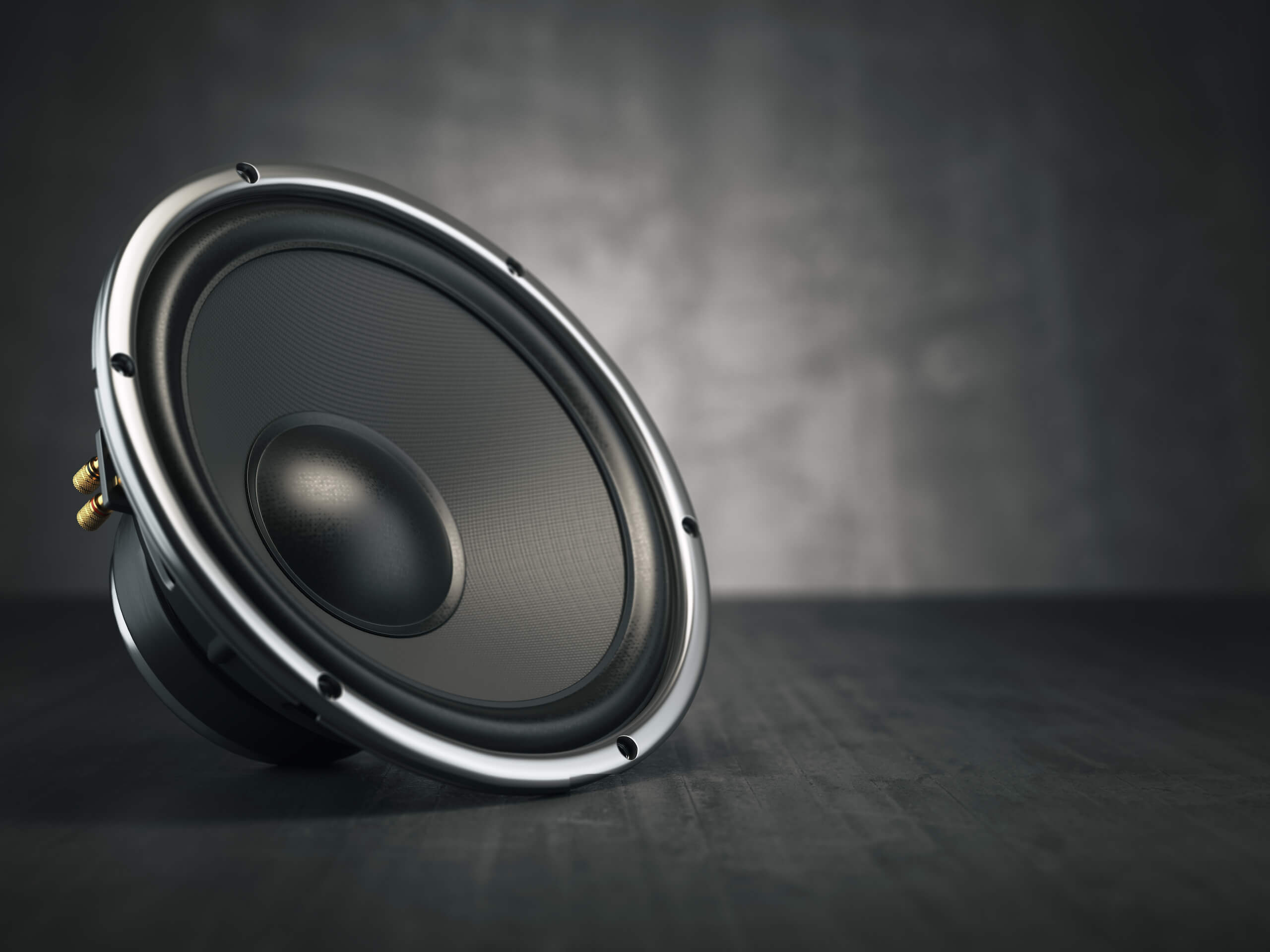 Close-up of a black speaker lying on its side against a dark, blurred background. The speaker cone is prominent, and the metallic outer edge is visible, with some screws around the perimeter. The surface beneath it is textured and dark, reminiscent of a TV wall mounting setup.