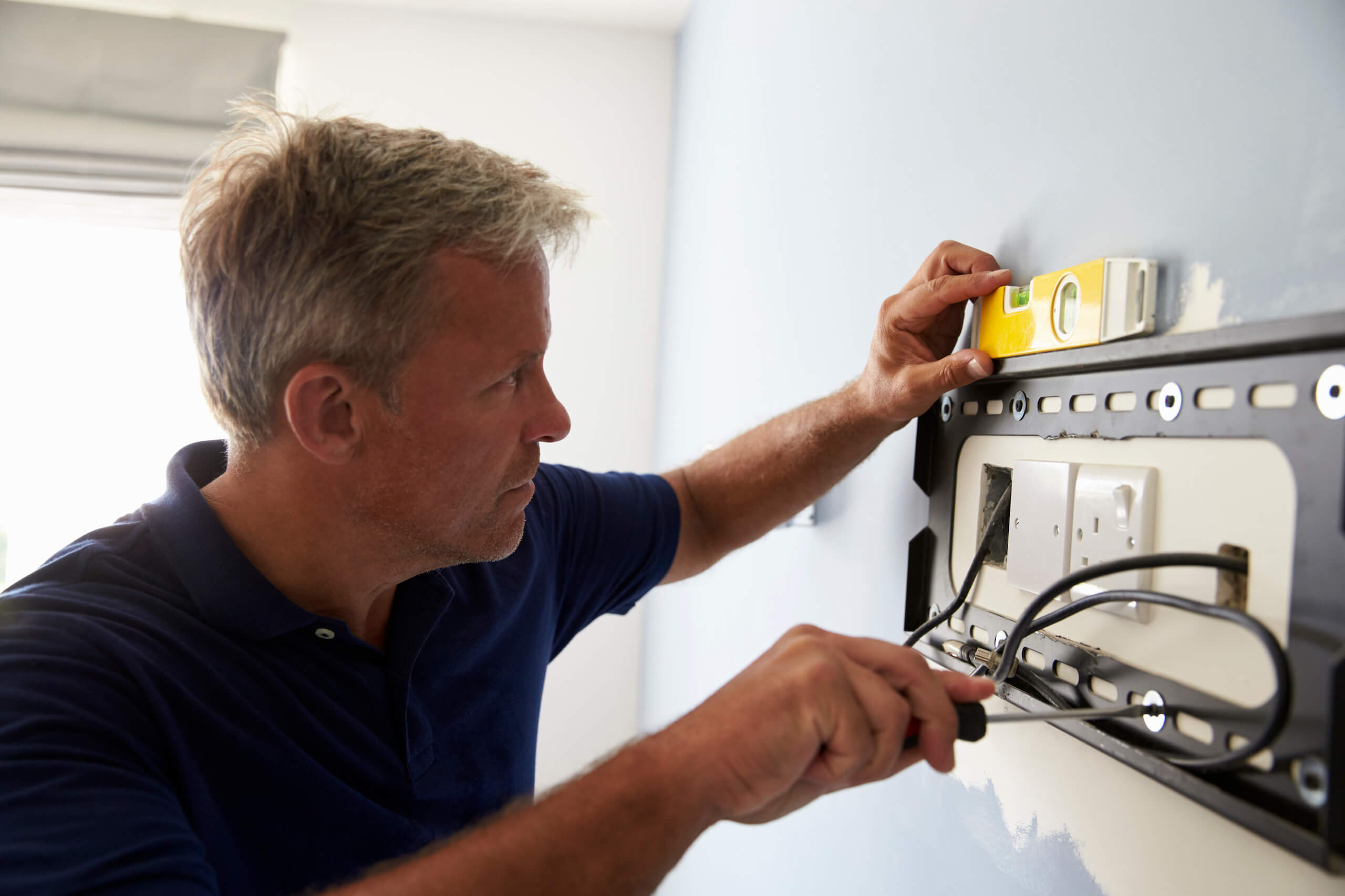 A man in a navy shirt uses a spirit level and screwdriver to install or adjust a TV wall mounting bracket. The surrounding area has a light blue wall with some paint patches. He is carefully aligning the unit to ensure it is level.
