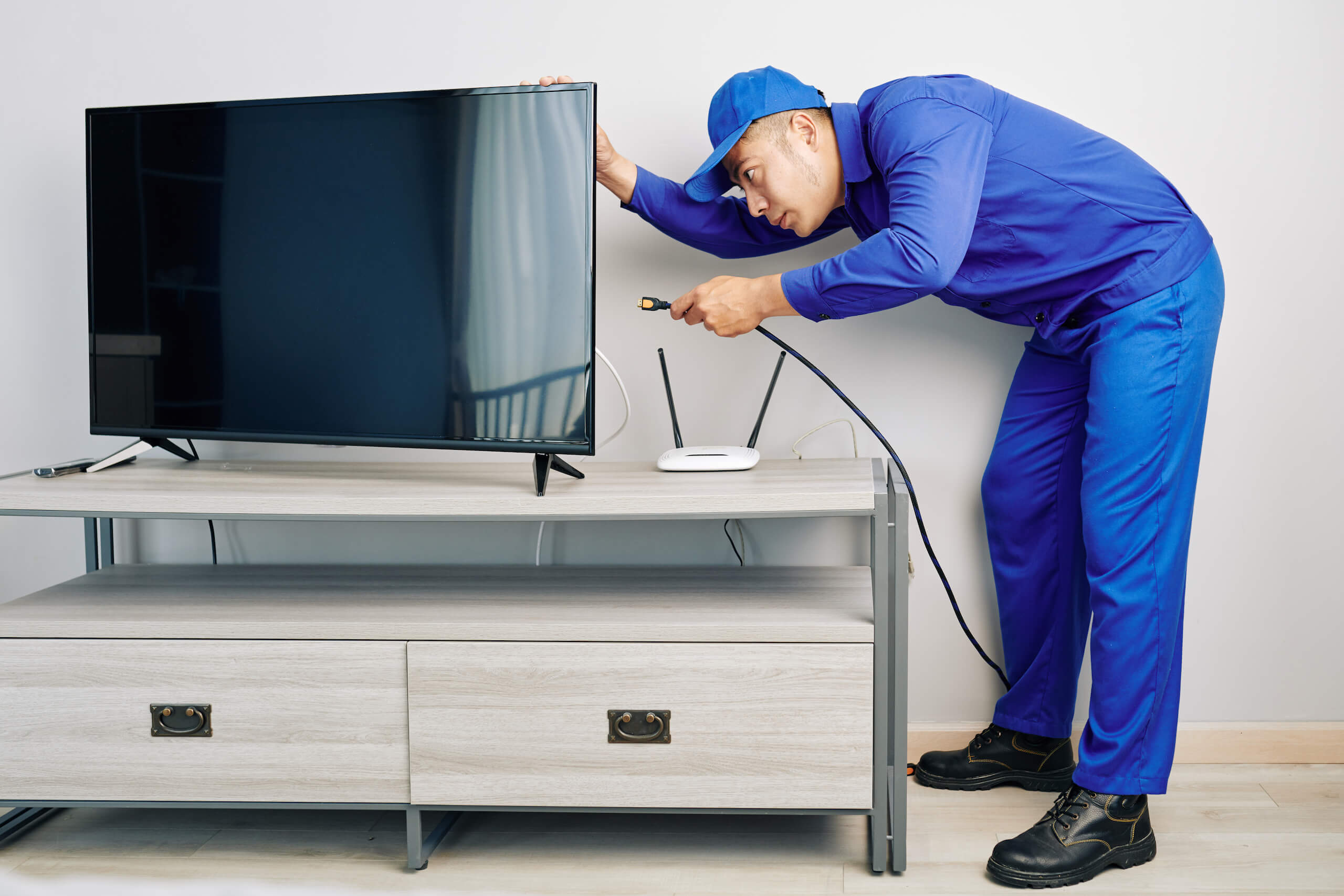 A technician in blue uniform and cap is setting up a television on a stand. He is holding an HDMI cable, appearing to connect it to the TV. A white Wi-Fi router sits on the stand next to the TV, though this setup seems temporary as TV wall mounting might be next. The background is a plain wall.