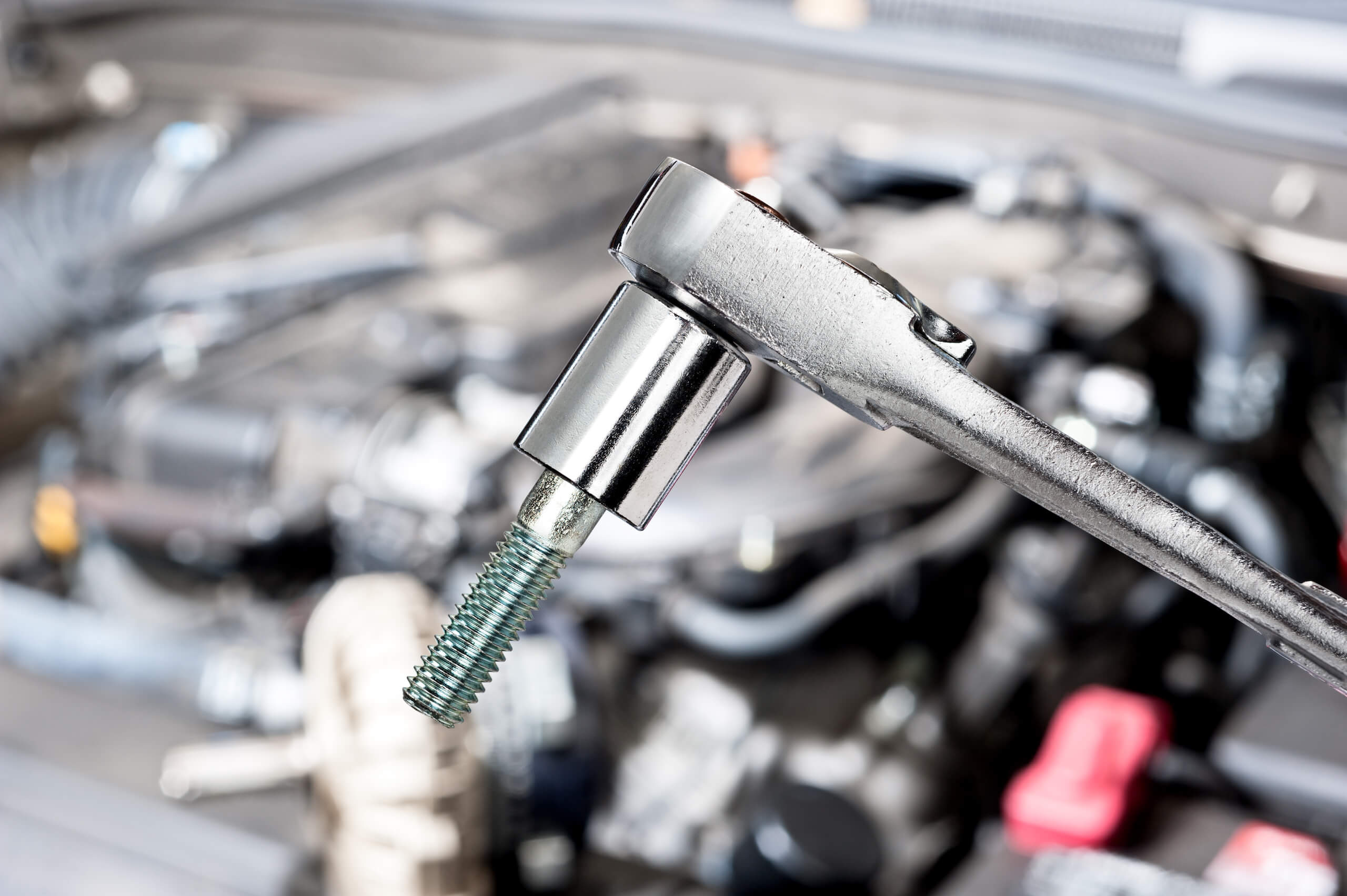 A close-up of a silver ratchet wrench holding a shiny bolt with a blue-green coating, reminiscent of those used in TV wall mounting. The background is blurred but appears to be the open hood of a vehicle, showing various engine components and parts.