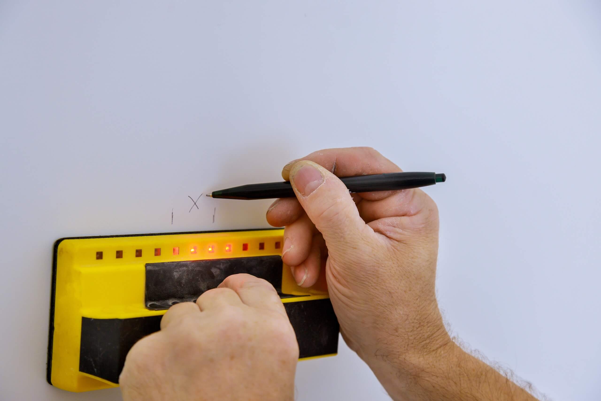 A person running a stud finder across a white wall and marking the stud edge in pencil before fixing a TV wall mount