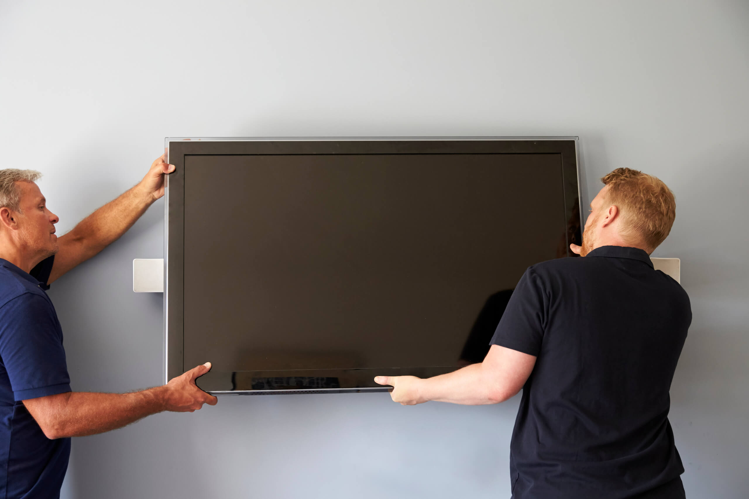 Two people are engaged in TV wall mounting, with one on the left side and the other on the right, carefully aligning and securing the flat-screen television in place. Both individuals appear focused on ensuring the TV is properly installed.