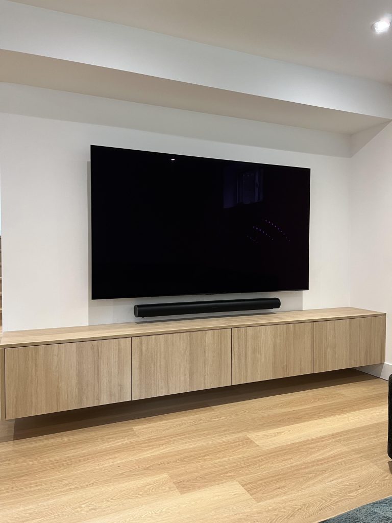Black soundbar mounted on the wall in the gap between a wall mounted TV and a floating oak cabinet