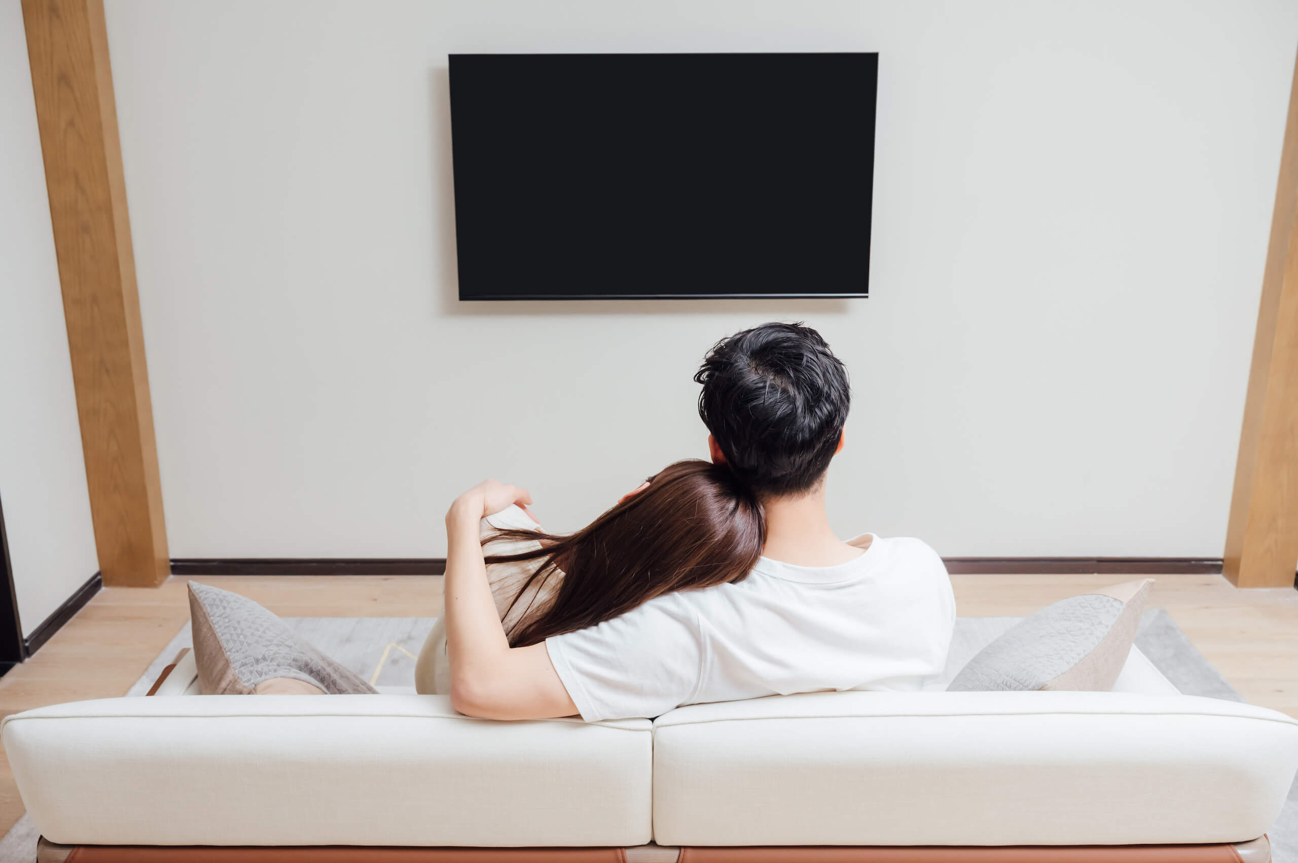 A couple on a white sofa, seen from behind, watching a TV mounted on the wall in front of them.