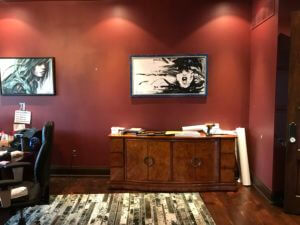 A room with dark red walls features two large paintings, one of a face in motion and another partially visible. There is a wooden sideboard with assorted items on it. A portion of a desk and chair is visible to the left, while a TV wall mounting sits above. A patterned rug covers the floor.