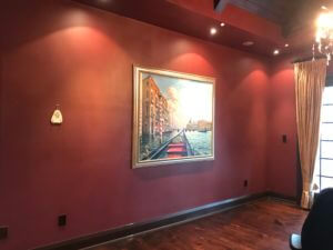 A room with maroon walls featuring a framed painting of a Venetian canal scene. The painting is centrally hung and depicts gondolas on water with buildings on both sides. The room, equipped for TV wall mounting, has hardwood flooring, recessed lighting, and a chandelier partially visible.