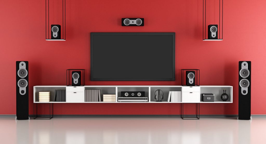 A modern home entertainment setup with a large flat-screen TV wall mounting on a red wall. Surrounding the TV, there are five black speakers: two tall floor-standing speakers, two smaller ones on shelves, and one center speaker above the TV. The shelves house books and decorative items.