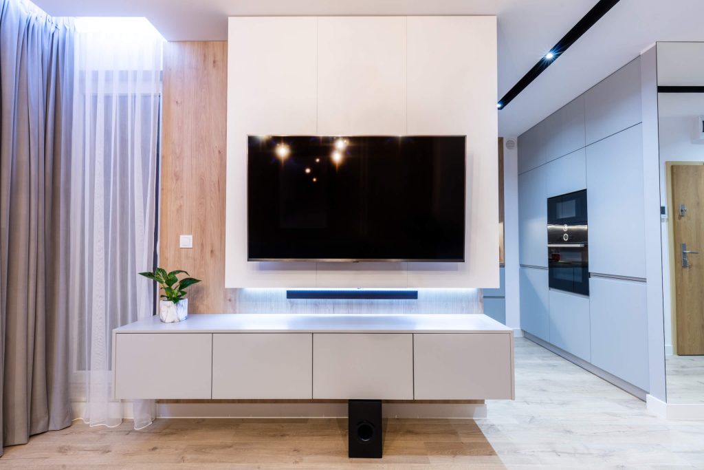 A modern living room showcases a large flat-screen TV with expert TV wall mounting above a sleek, white media console. A soundbar is placed below the TV, with a small plant on the left side of the console. The space is bright, featuring light wood accents and floor.