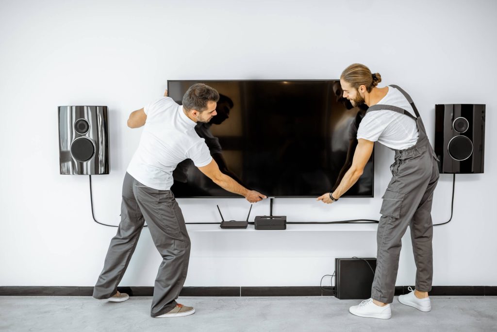 Two men are mounting a flat-screen TV on a white wall. They are wearing white shirts and gray overalls. The TV, expertly installed using a TV wall mounting system, is centered between two wall-mounted speakers, with a shelf below holding a router and other electronic devices. Both men are adjusting the TV's position.